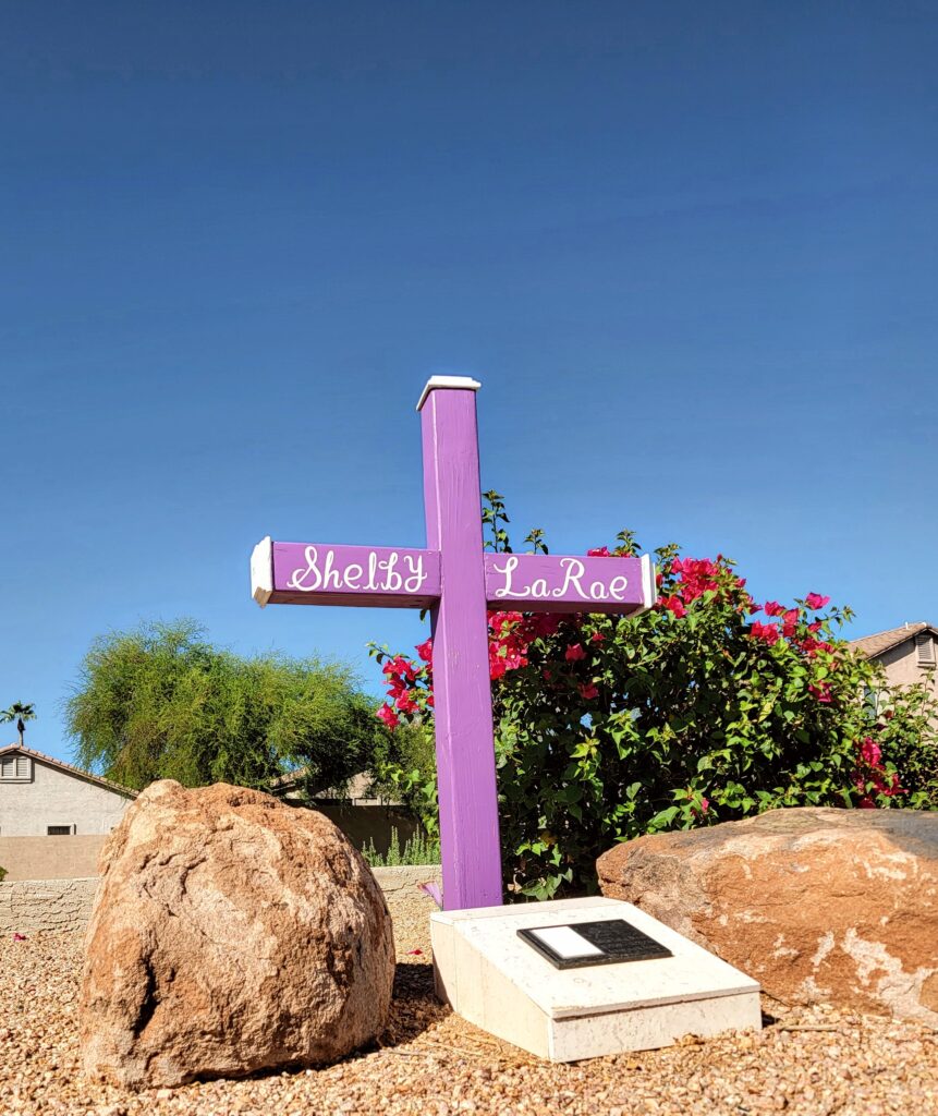 Purple memorial cross reading “Shelby LaRae” set in desert gravel between two large boulders, with blooming bougainvillea behind it.