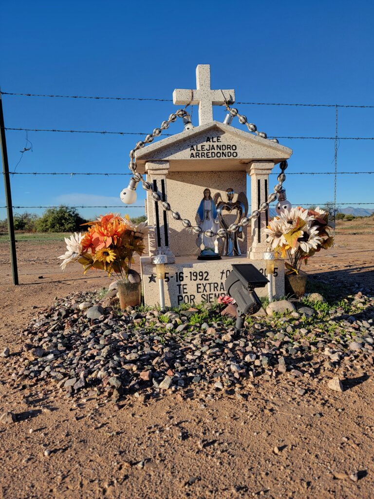 Roadside memorial shrine for Alejandro Arredondo with a stone cross, flowers, and Virgin Mary statue in the Arizona desert.