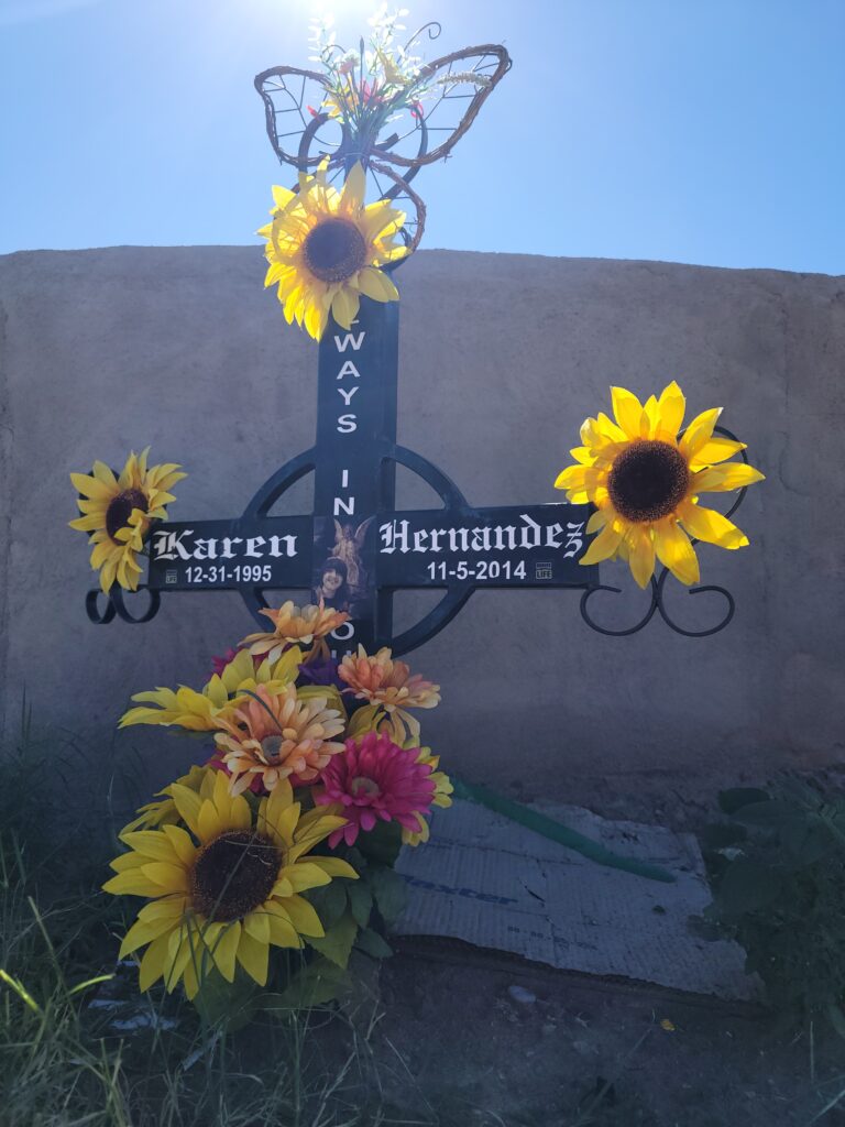 Sunflower-covered roadside memorial cross for Karen Hernandez (12-31-1995 to 11-5-2014) beside a stucco wall.