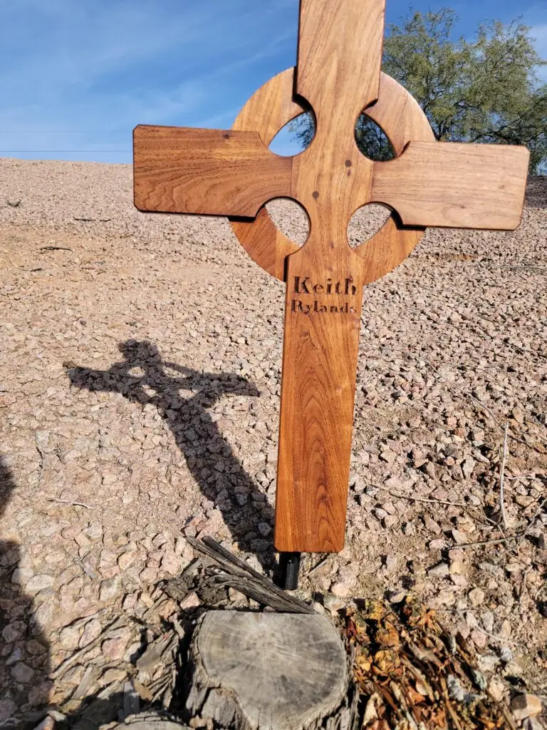 Wooden Celtic-style cross memorial for Keith Rylands on a rocky desert roadside, casting a shadow in bright sun.