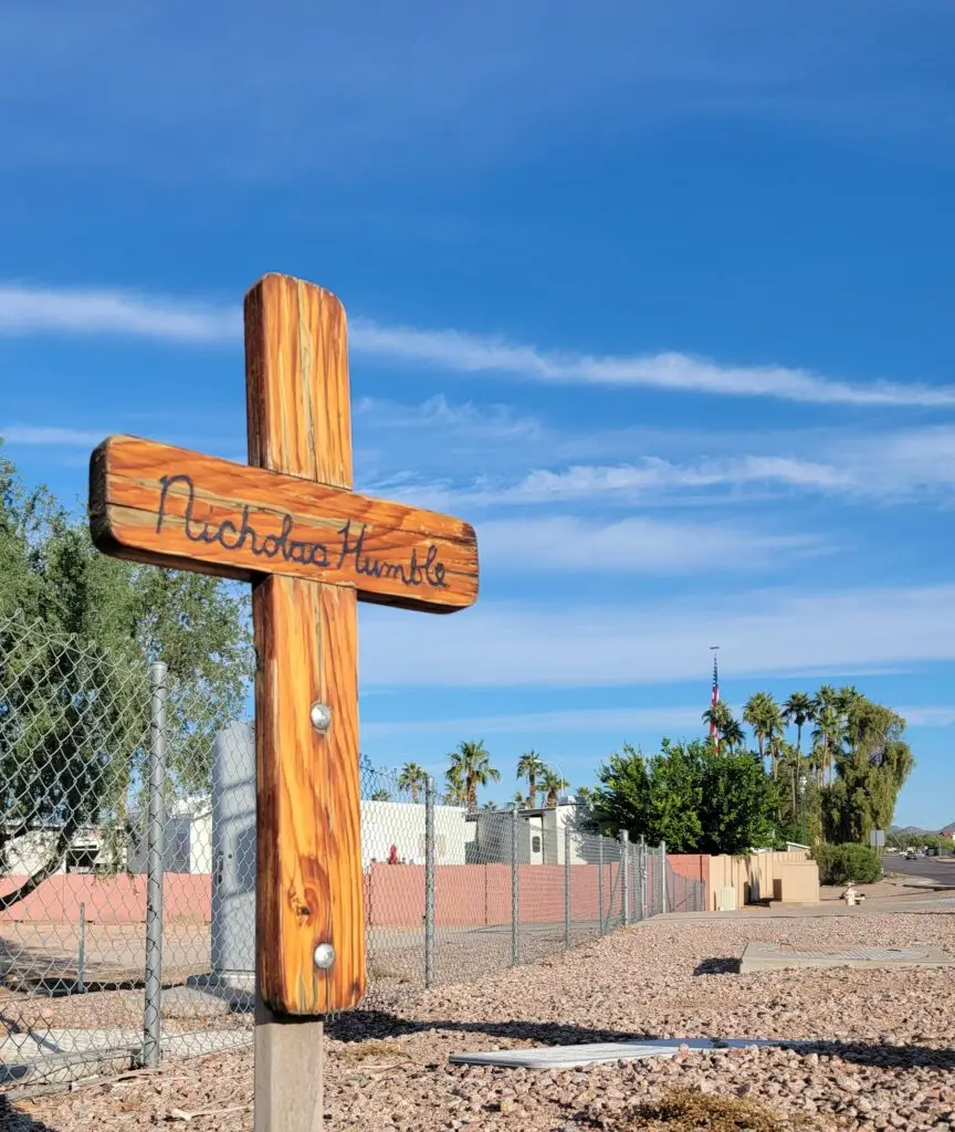 Wooden roadside memorial cross reading “Nicholas Humble” beside a chain-link fence under a bright blue sky.